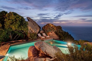 Une piscine entourée de grands rochers et d’arbres, avec des chaises longues, et offrant une vue sur l’océan.