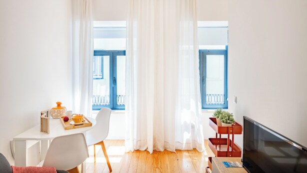 Une pièce lumineuse avec une table blanche, une chaise, un téléviseur et une fenêtre avec des volets bleus.