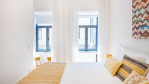 Une pièce lumineuse et blanche, avec un lit, un tapis tissé et une vue sur un balcon à travers de fines rideaux.