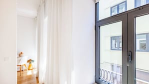 Une pièce lumineuse avec une grande fenêtre, un plancher en bois, une chaise blanche et une petite table sur laquelle est posé un vase.