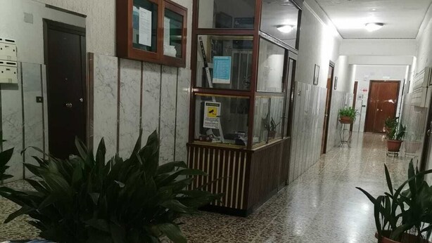 Un hall d’entrée doté d’un sol en terrazzo, un comptoir de réception protégé par une paroi de verre et des plantes en pot.