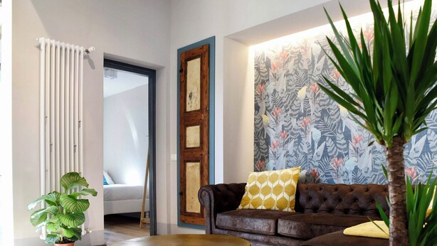 Una sala de estar con un sofá marrón, un otoman redondo, una planta en maceta y una pared con diseño.