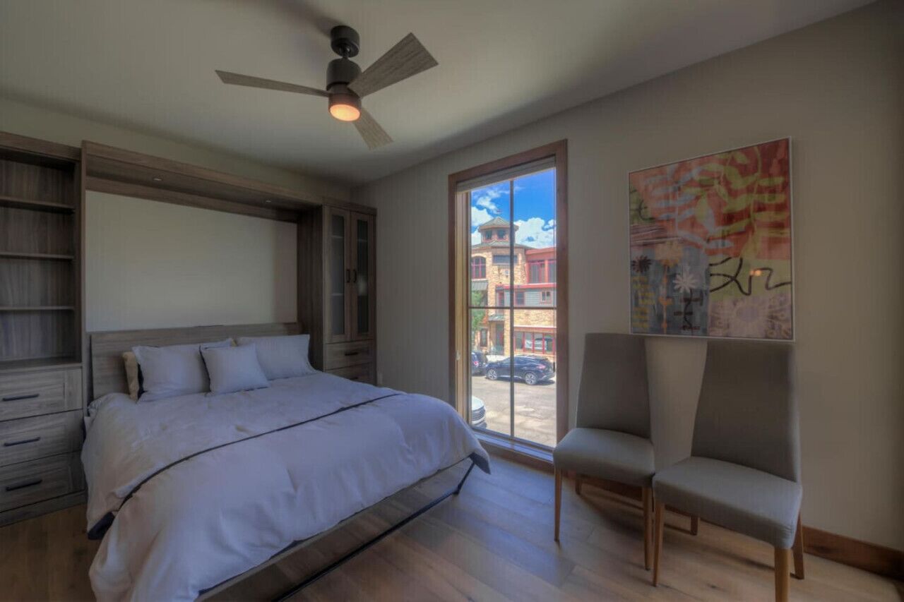 Un dormitorio con una cama grande, un ventilador de techo, una ventana con vistas a edificios y una silla.