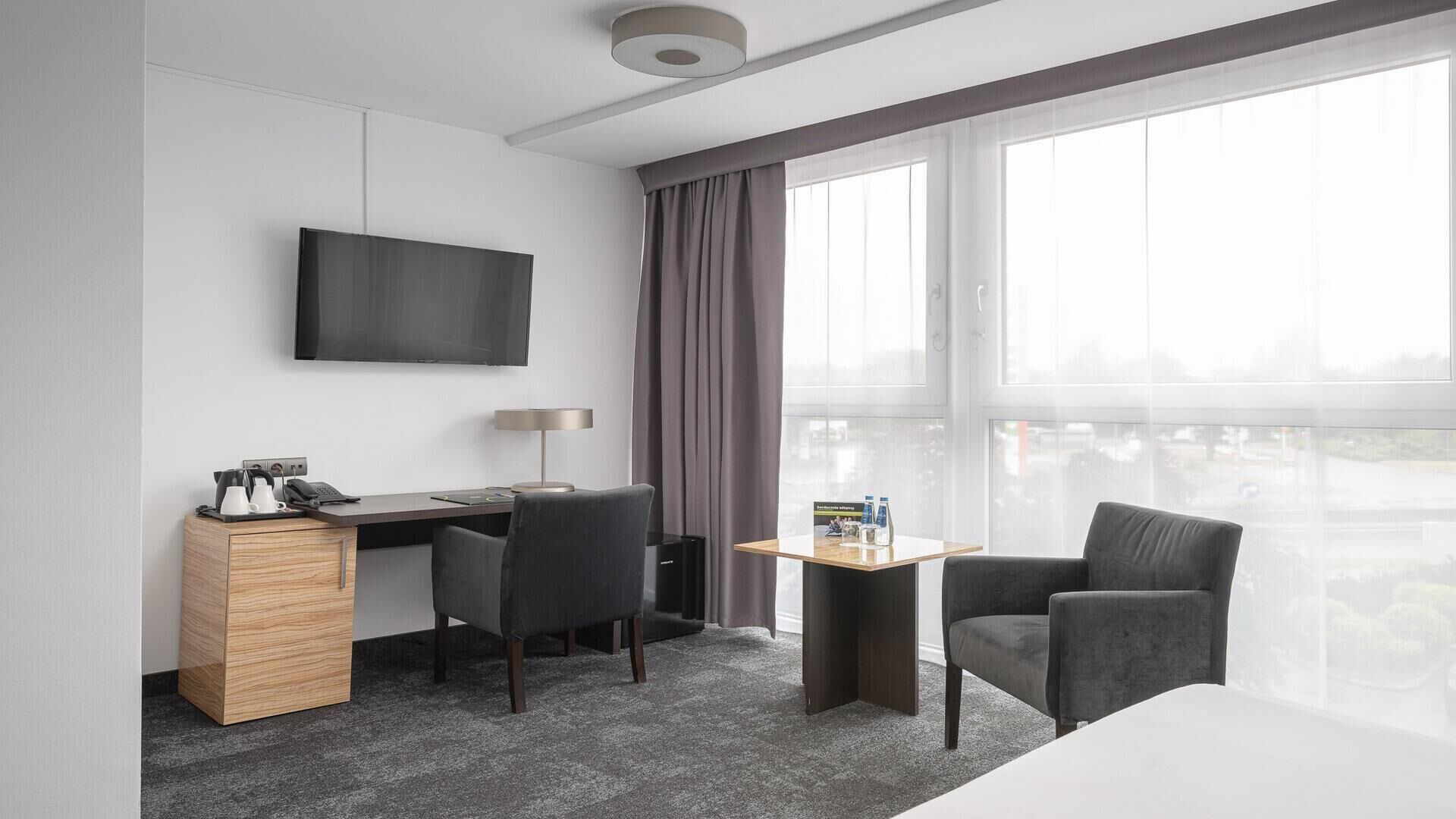 Ein modernes Hotelzimmer mit Fernseher, Schreibtisch mit Lampe, einem kleinen Tisch mit Wasserflaschen, zwei Stühlen und einem großen Fenster mit Blick auf die Stadt.
