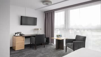 Ein modernes Hotelzimmer mit Fernseher, Schreibtisch mit Lampe, einem kleinen Tisch mit Wasserflaschen, zwei Stühlen und einem großen Fenster mit Blick auf die Stadt.