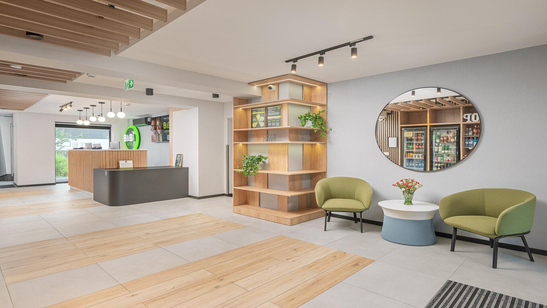 Un hall d’accueil moderne avec un comptoir en bois, un grand miroir rond et deux fauteuils verts.