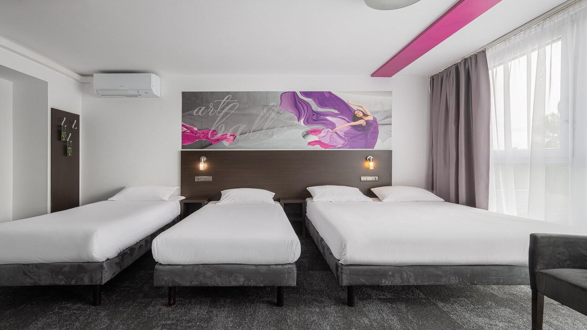 Habitación de hotel con tres camas individuales, un mural grande en la pared, dos lámparas de noche, una ventana con cortinas y una lámpara en el techo.