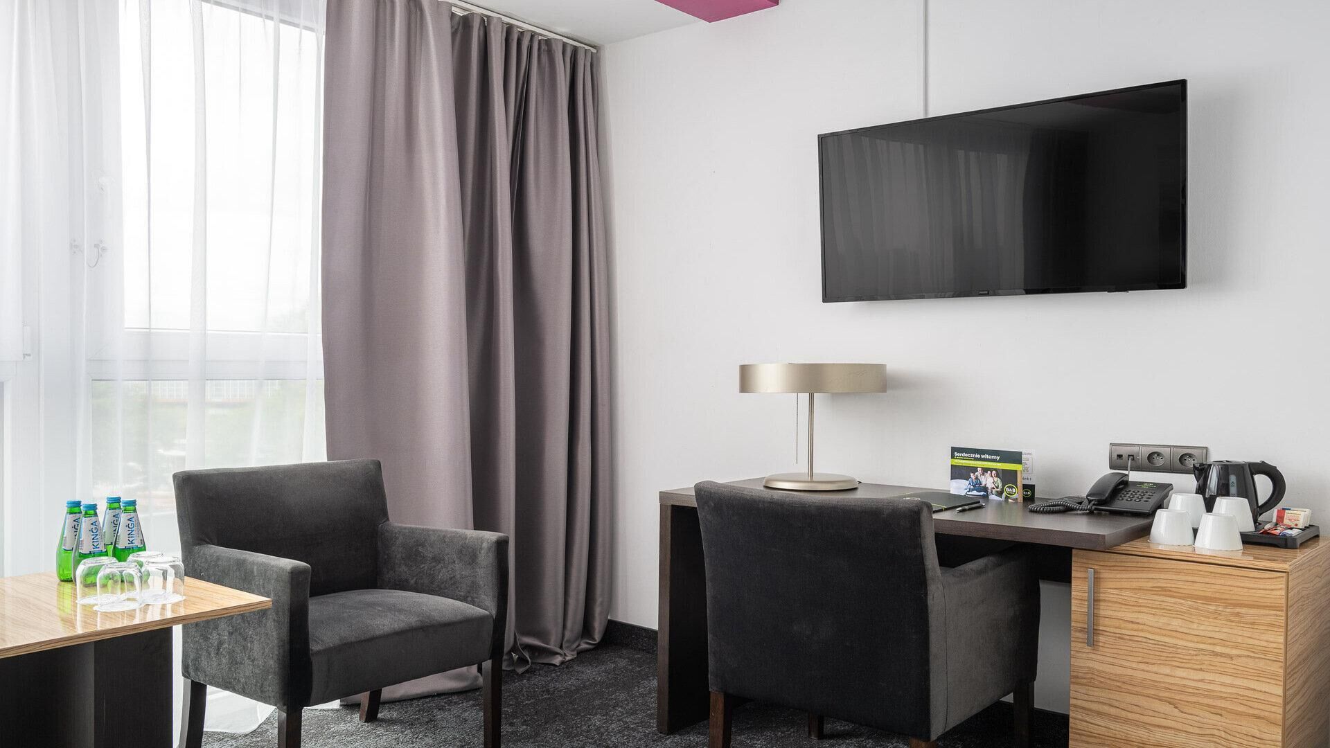 Une chambre d’hôtel équipée d’un bureau, de deux chaises, d’une télévision, d’une lampe, d’une machine à café et d’un mini-bar.