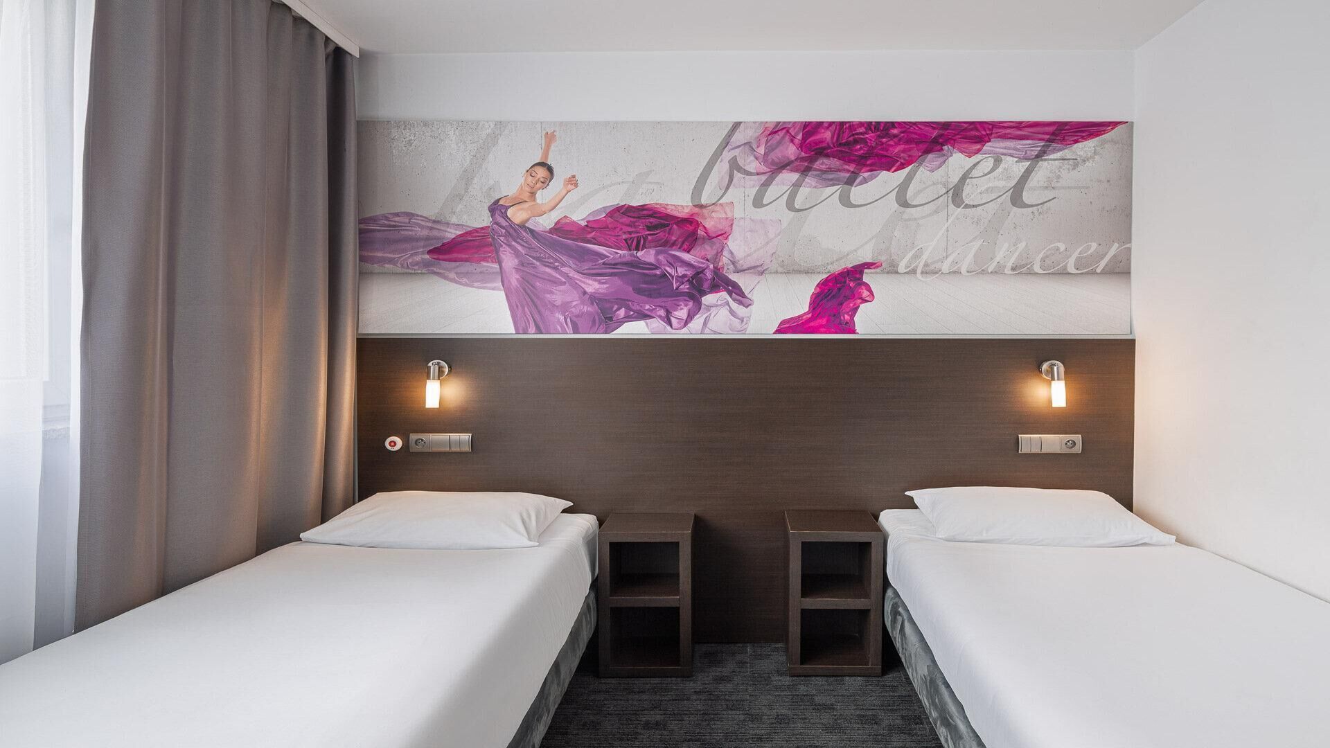 Hotellihuone, jossa on kaksi erillistä sänkyä, suuri seinämaalaus ja kaksi seinään kiinnitettyä valaisinta.