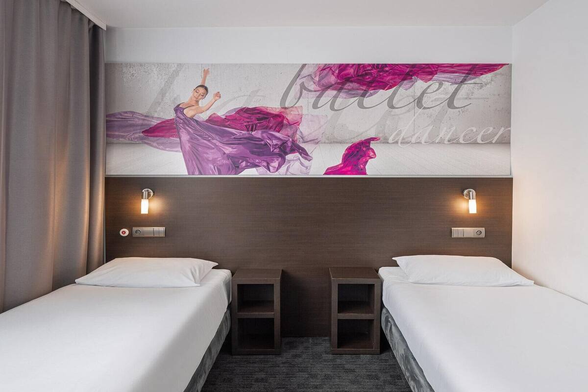 A hotel room with two single beds, a large wall mural, and two wall-mounted light fixtures.