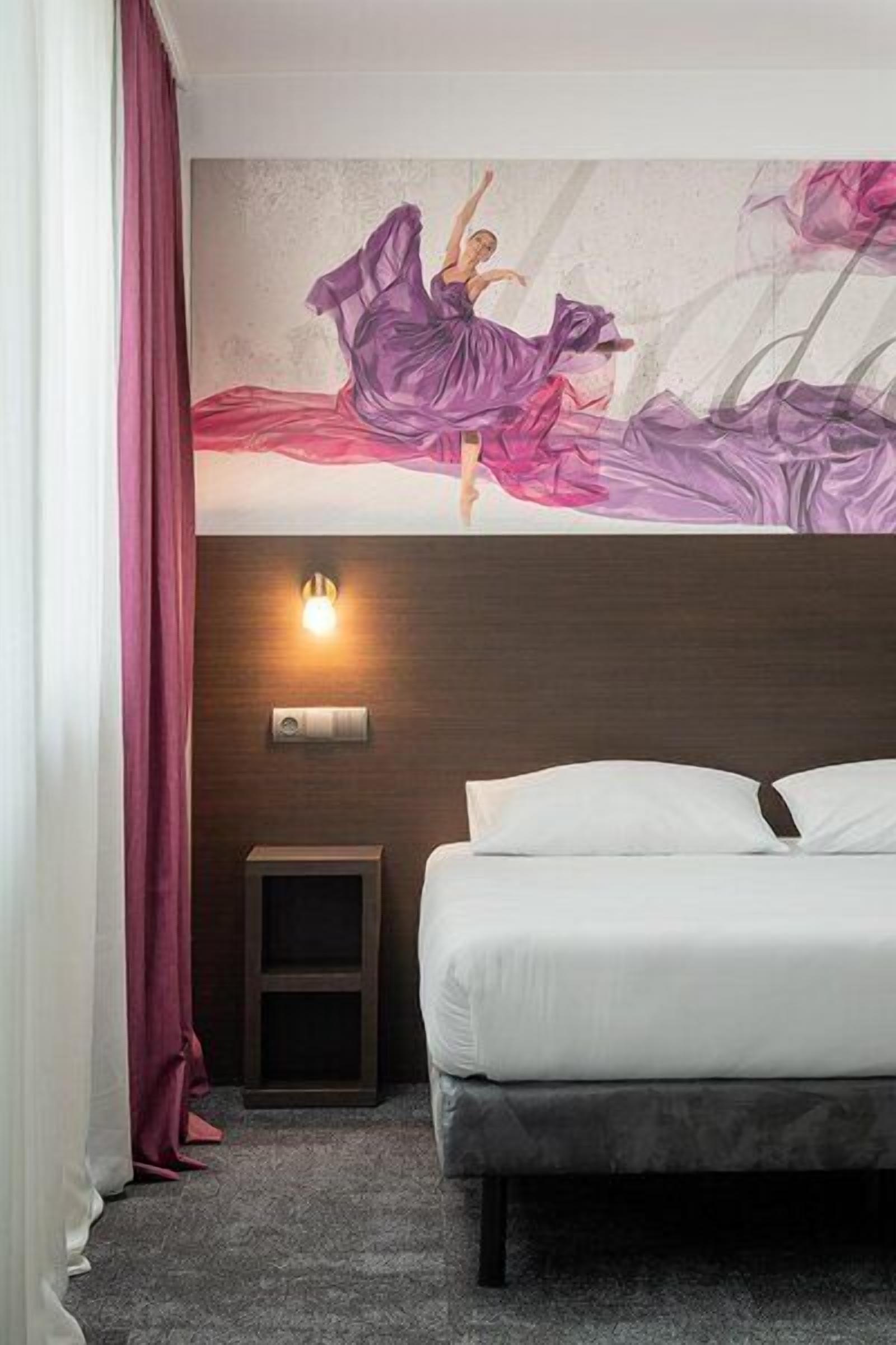 Habitación de hotel con cama, mesita de noche, lámpara de pared y un mural decorativo.