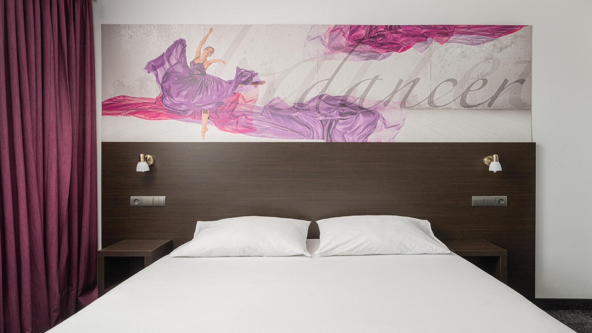 Une chambre d’hôtel comprenant un lit, deux tables de chevet, une petite table, un luminaire fixé au mur et un grand tableau représentant une danseuse.