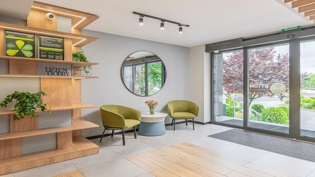A modern reception area with a wooden reception desk, two green chairs, a round mirror, and a view of the outdoors through large glass doors. - B&B HOTEL Bytom (Bytom)