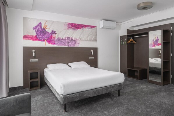 A hotel room with a bed, a wardrobe, a mirror, and a large abstract painting on the wall.