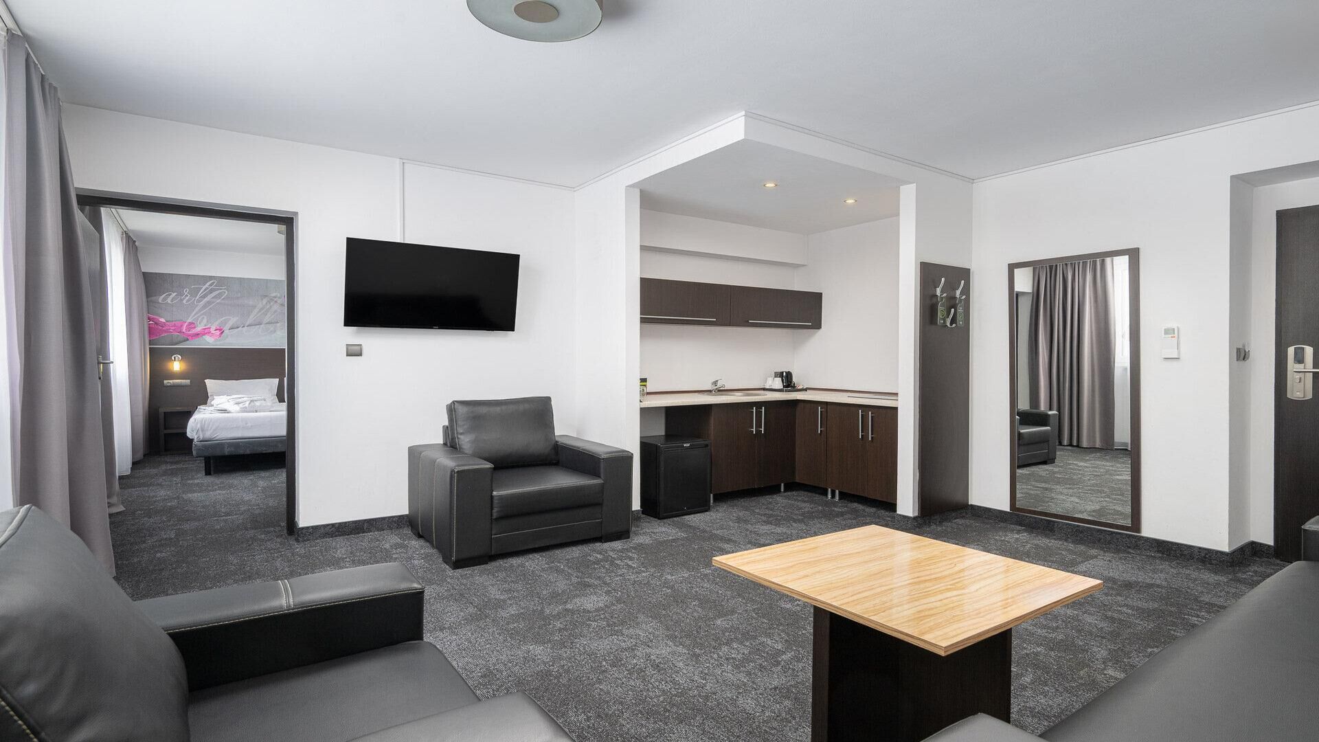 Una habitación de hotel moderna con sofá, una mesita, televisor, kitchenette y baño a la vista a través de una puerta abierta.