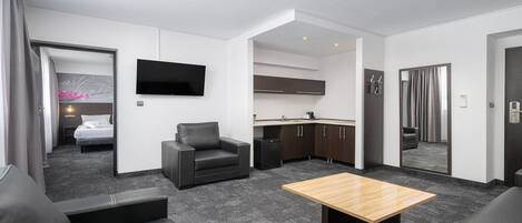 Una habitación de hotel moderna con sofá, una mesita, televisor, kitchenette y baño a la vista a través de una puerta abierta.