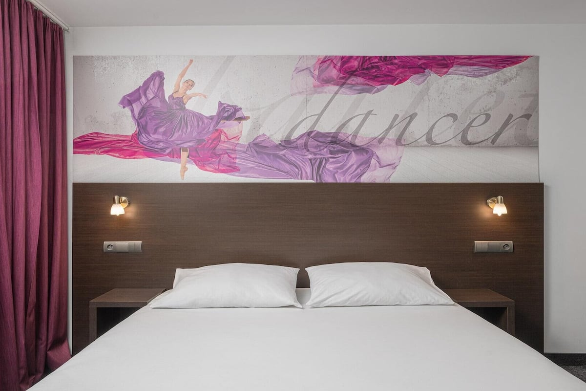 A hotel room with a bed, two bedside tables, a headboard with wall sconces, and a wall mural with the word "dancer" and a figure in a purple dress.