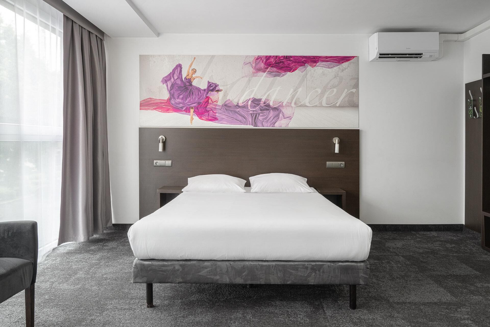 A hotel room with a bed, a headboard with a control panel, a wall-mounted air conditioner, and a window with curtains.