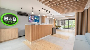 A modern hotel reception area with a wooden reception desk, a large B&B Hotels sign, a flat-screen TV, and a seating area with a view of the outdoors. - B&B HOTEL Bytom (Bytom)