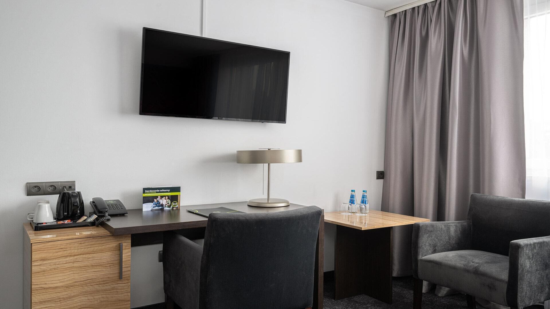 A hotel room with a flat-screen TV mounted on the wall, a desk with a lamp, two chairs, a small table with water bottles, and a minibar.