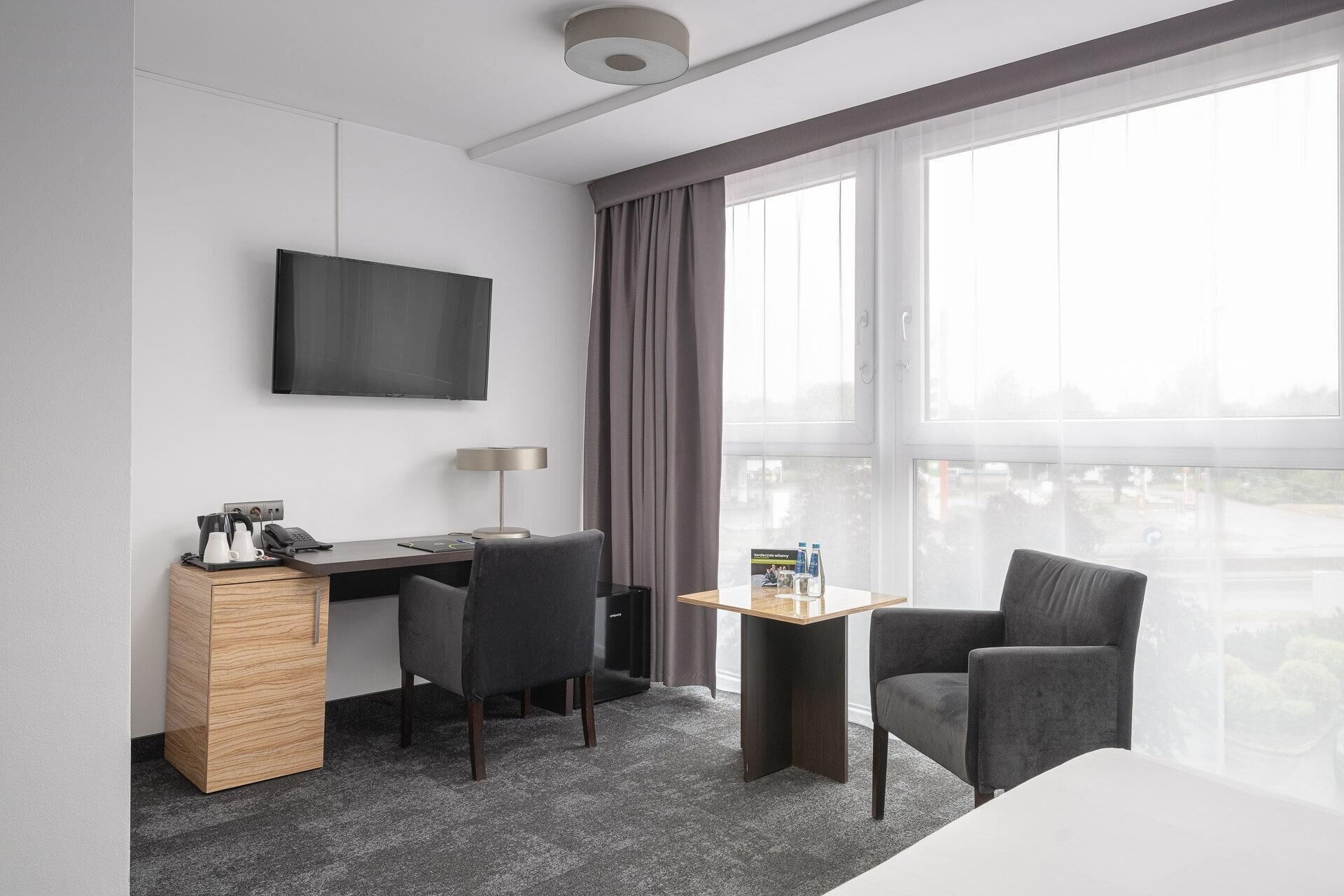 A hotel room with a desk, two chairs, a TV, a small table with a bottle, and a window with curtains.