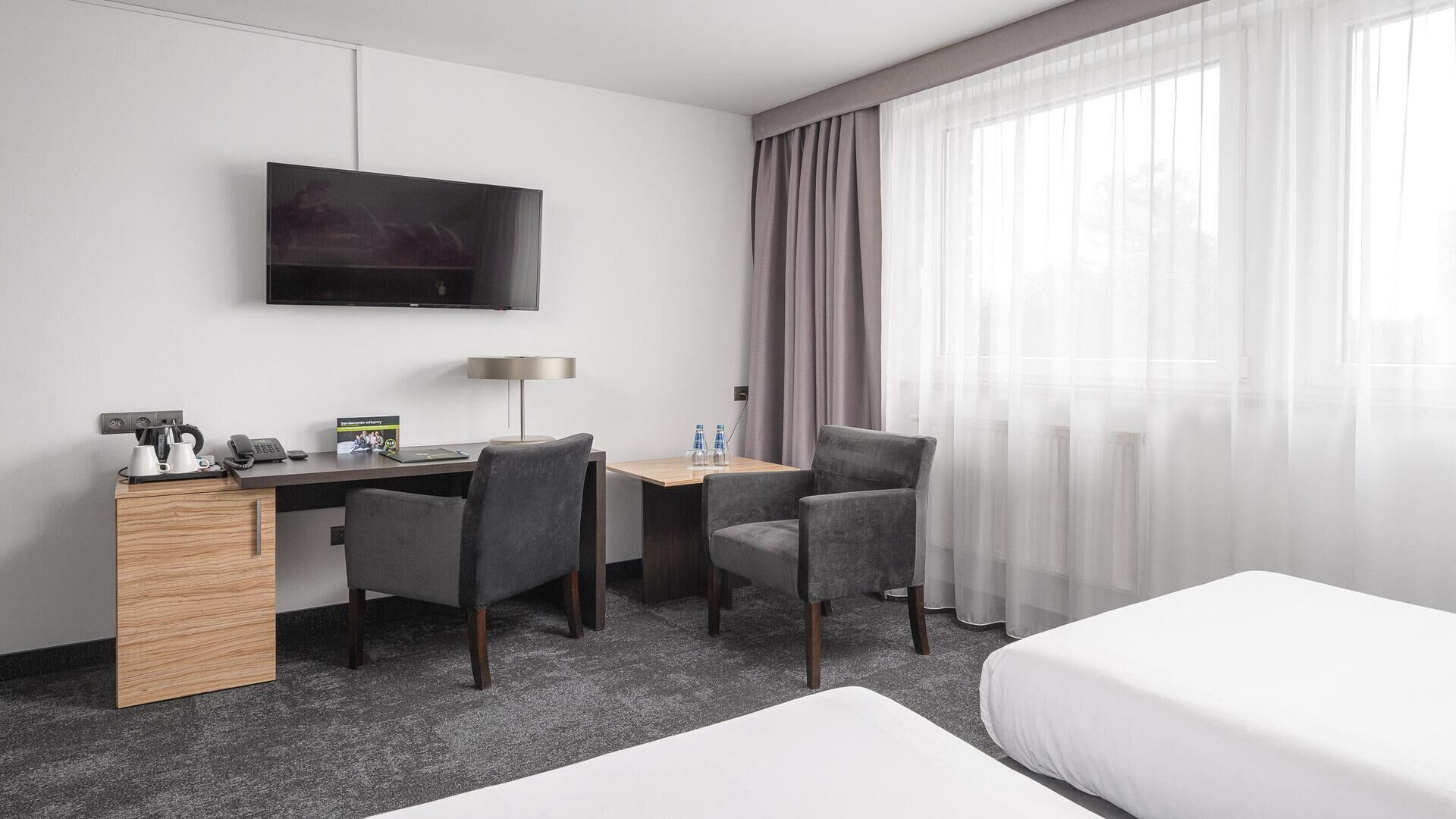 A hotel room with a desk, two chairs, a bed, a TV, and a window with sheer curtains.