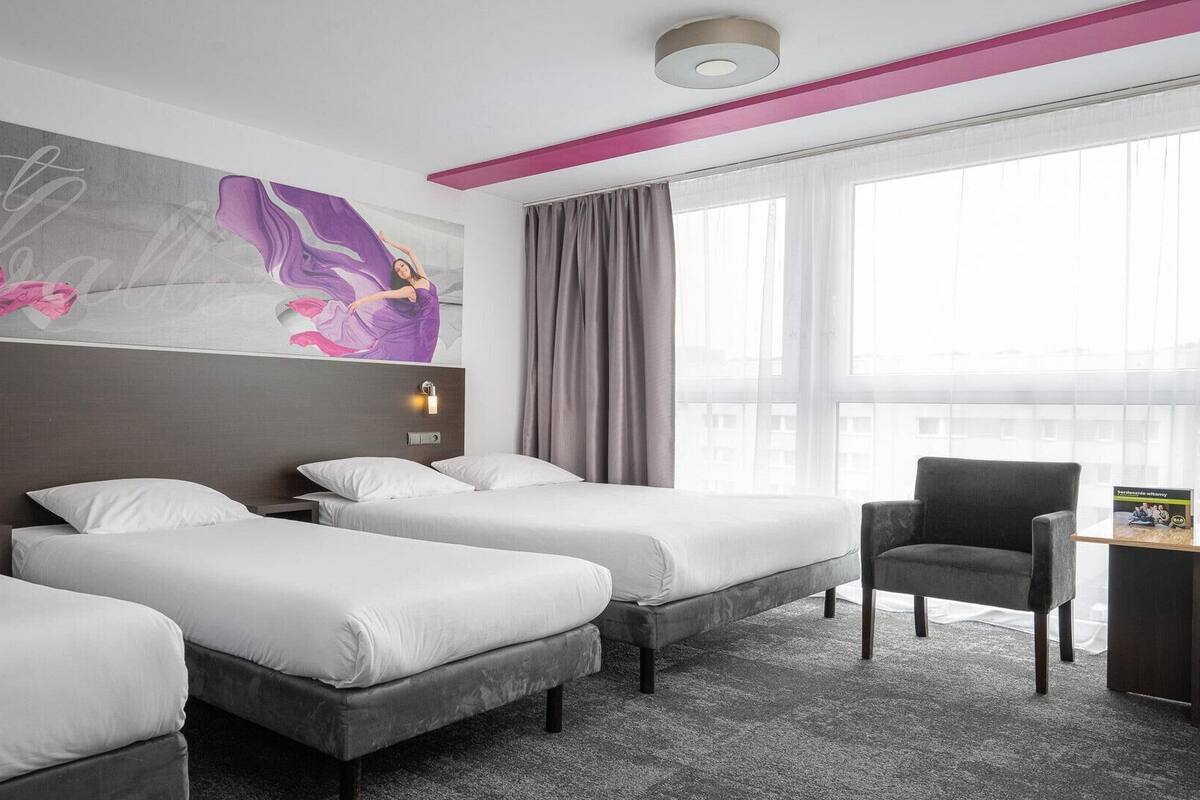 A hotel room with three beds, a desk, and a chair. There is a large window with curtains, a ceiling light, and a wall with a floral artwork.