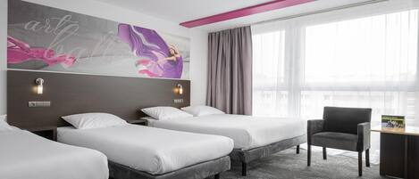 Habitación de hotel con tres camas, un escritorio y una silla. Cuenta con un ventanal con cortinas, una lámpara en el techo y un cuadro floral en la pared.