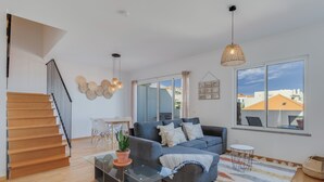 A modern living room with a sofa, a glass coffee table, a potted plant, a staircase, and large windows offering a view of buildings. - Alta Garajau I (Caniço)