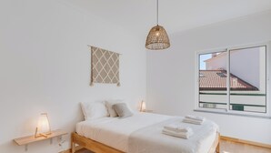 A minimalist bedroom with a wooden bed, bedside lamps, a hanging light fixture, a window with a view of a building, and a wall-mounted decorative piece. - Alta Garajau I (Caniço)