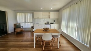 A modern kitchen with white cabinets, a wooden dining table with four chairs, and a large window with blinds. - Guest Suite Bredbo (Jerangle)