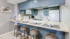 A modern kitchen with teal cabinets, a white countertop, and three woven stools. - Amelia South F1 - Hosted by Moore AI Rentals (Fernandina Beach)