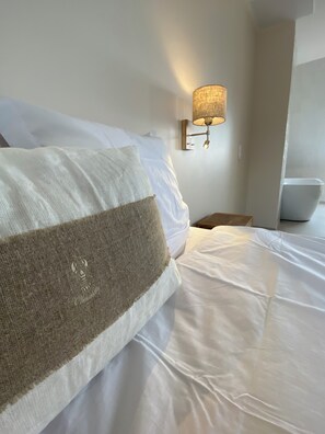 Uma cama bem arrumada com roupa de cama branca e travesseiro marrom, uma luminária na parede e uma pequena mesa de cabeceira de madeira.