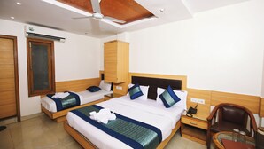 A hotel room with two beds, wooden headboards, a ceiling fan, an air conditioning unit, a desk with a chair, and a small table with a glass top. - Hotel Lav Kush Deluxe (New Delhi)