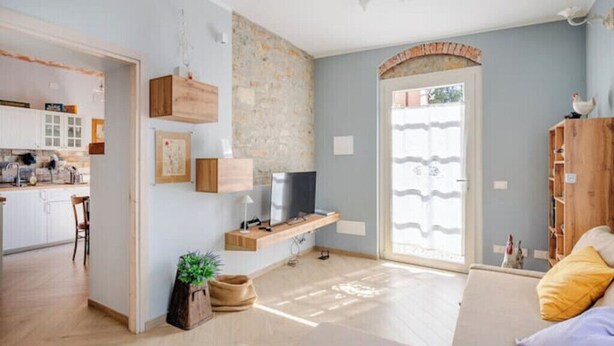 A modern living room with a sofa, a TV on a wooden stand, a kitchen in the background, and a stone wall.