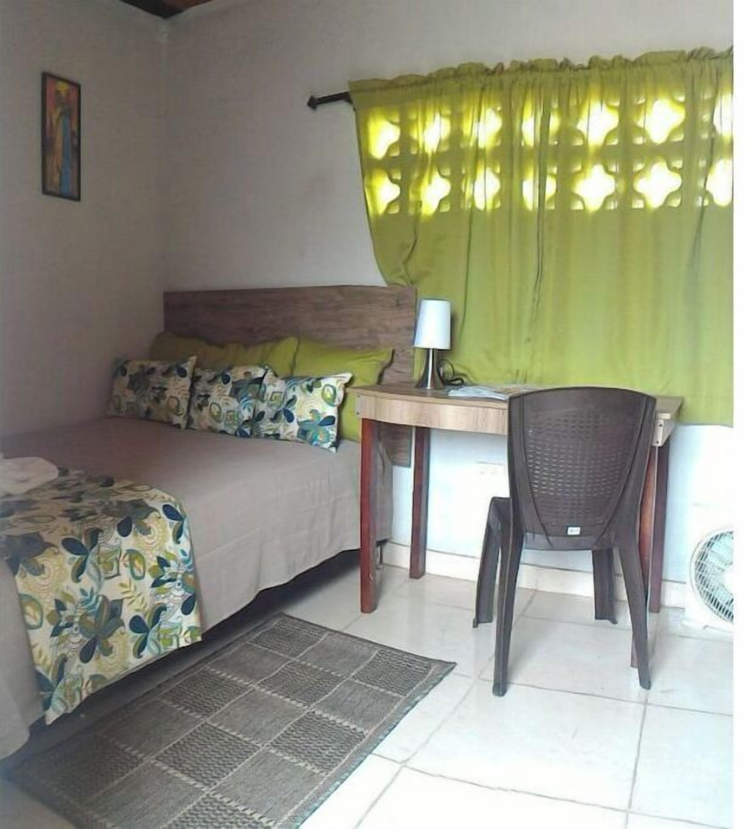 A small, well-lit room with a bed, a desk, and a chair. There is a window with green curtains, a wall-mounted lamp, and a small fan.