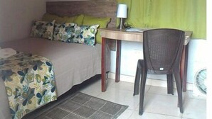 A small, well-lit room with a bed, a desk, and a chair. There is a window with green curtains, a wall-mounted lamp, and a small fan. - Yopal Apartment (Medellín)