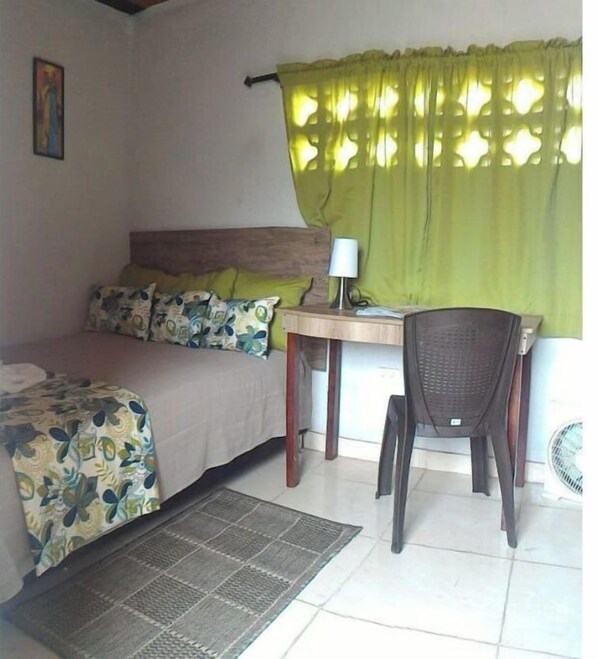 A small, well-lit room with a bed, a desk, and a chair. There is a window with green curtains, a wall-mounted lamp, and a small fan.