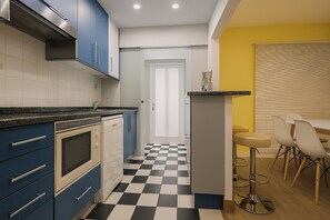 Una cocina con armarios azules, piso a cuadros blanco y negro, una puerta blanca y una pared amarilla con persianas.