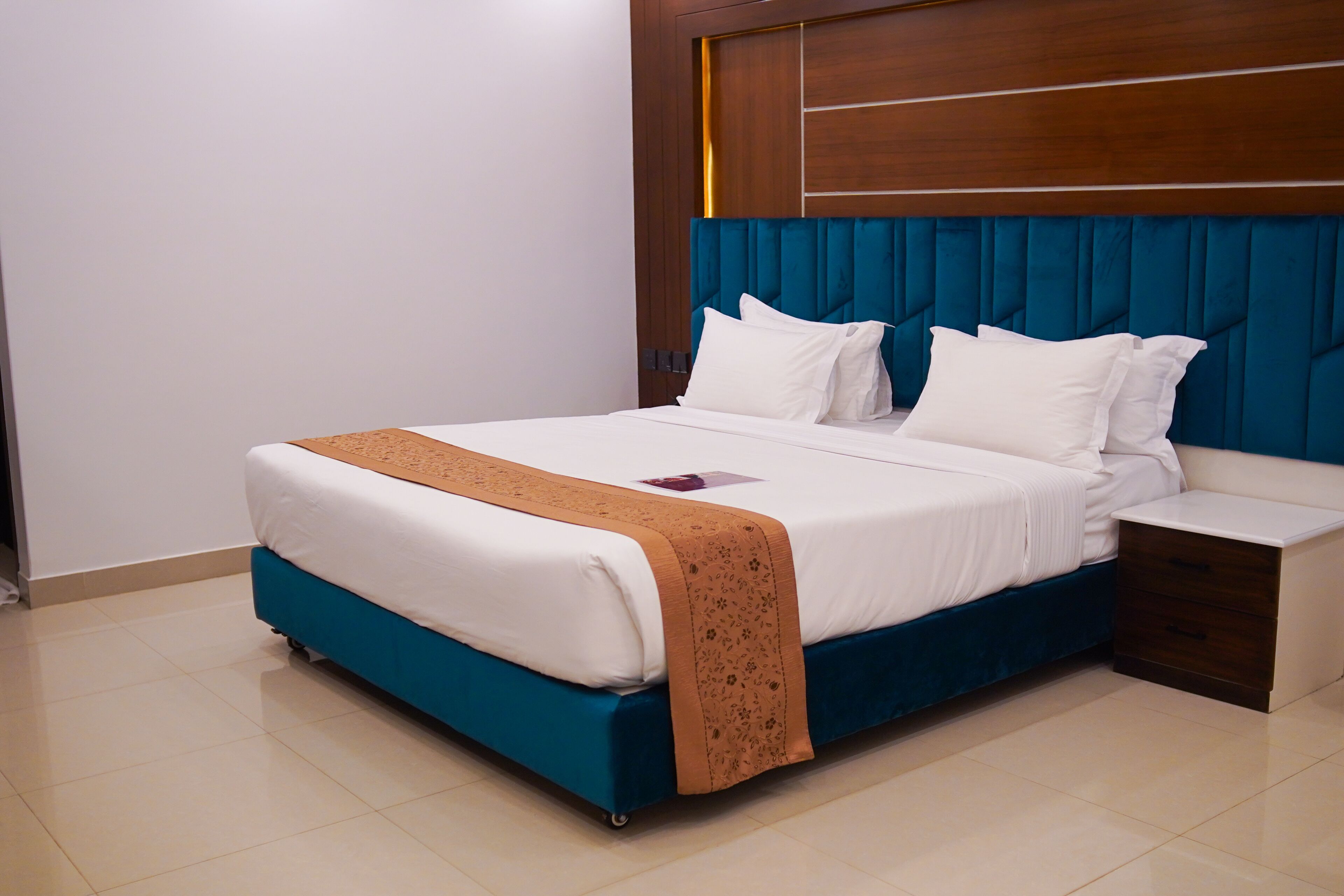 A neatly made bed with a blue headboard and white bedding, a bedside table with drawers, and a clean, tiled floor.