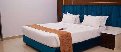 A neatly made bed with a blue headboard and white bedding, a bedside table with drawers, and a clean, tiled floor.