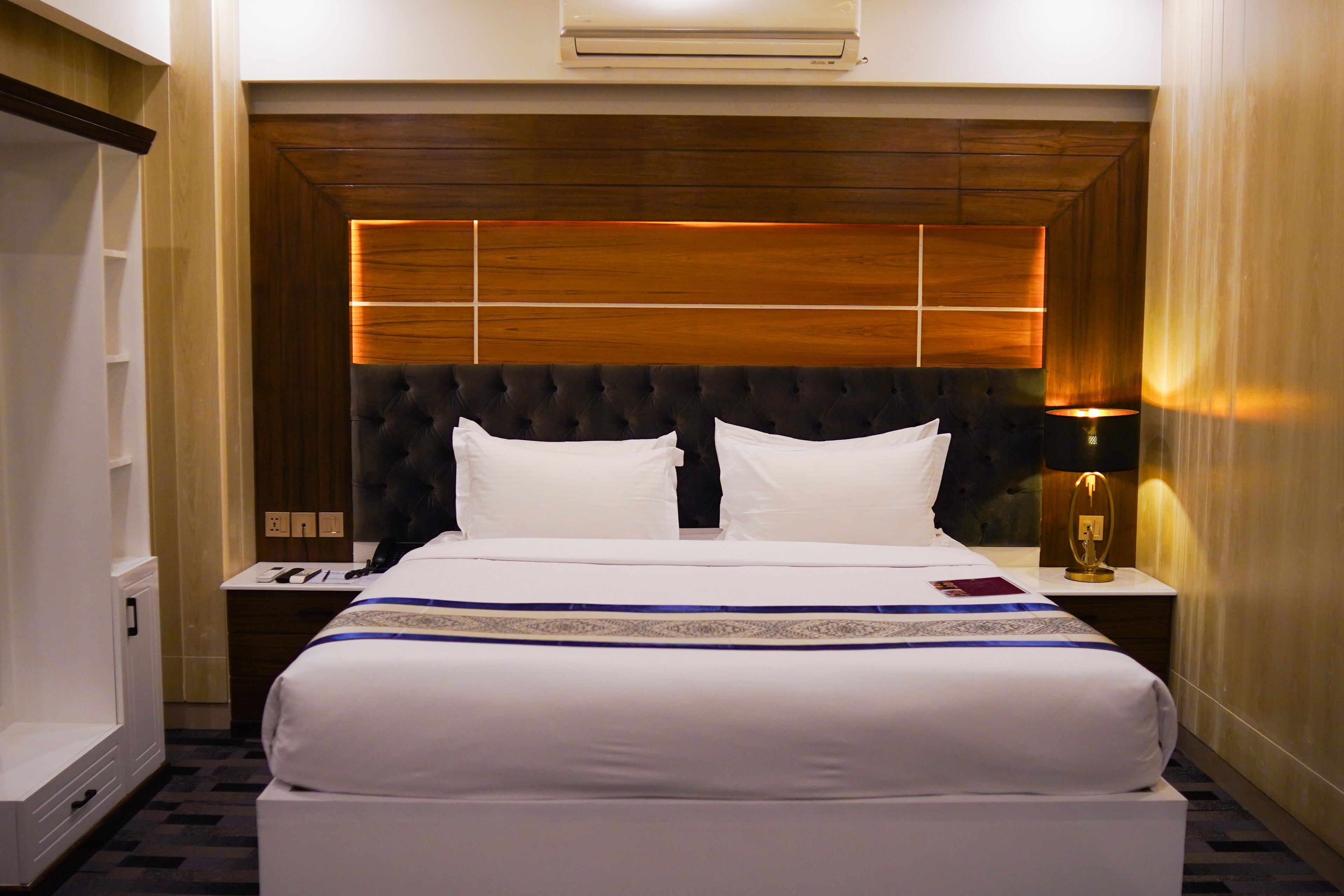 Ein Hotelzimmer mit einem großen Bett, einem hölzernen Kopfteil, einem Nachttisch mit Lampe und einer Wand-Klimaanlage.