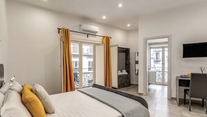 Un dormitorio con cama, escritorio y televisor. Tiene un balcón con cortinas amarillas y vistas a edificios.