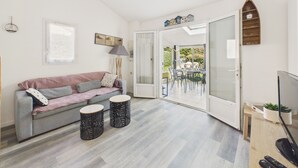 Un salon moderne avec un canapé gris, deux petites tables d’appoint rondes, un téléviseur et une porte-fenêtre coulissante donnant sur un espace repas extérieur.