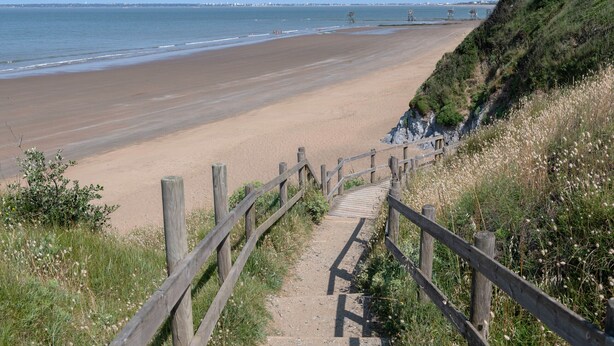 Un escalier en bois mène à une plage de sable, d’où l’on a une vue dégagée sur l’océan et les bâtiments situés au loin.
