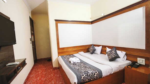 A hotel room with a wooden bed, a bedside table with a phone, a TV on a stand, and a red carpeted floor. - Hotel Jindal  (New Delhi)