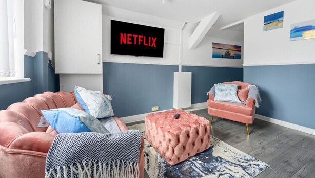 Un soggiorno moderno con un divano rosa, un poggiapiedi rosa e una poltrona rosa. Un televisore a schermo piatto montato a parete mostra il logo di Netflix.