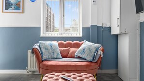 Una stanza con un divano rosa, un telecomando su un pouf abbinato, una finestra con grate e un quadro appeso al muro.