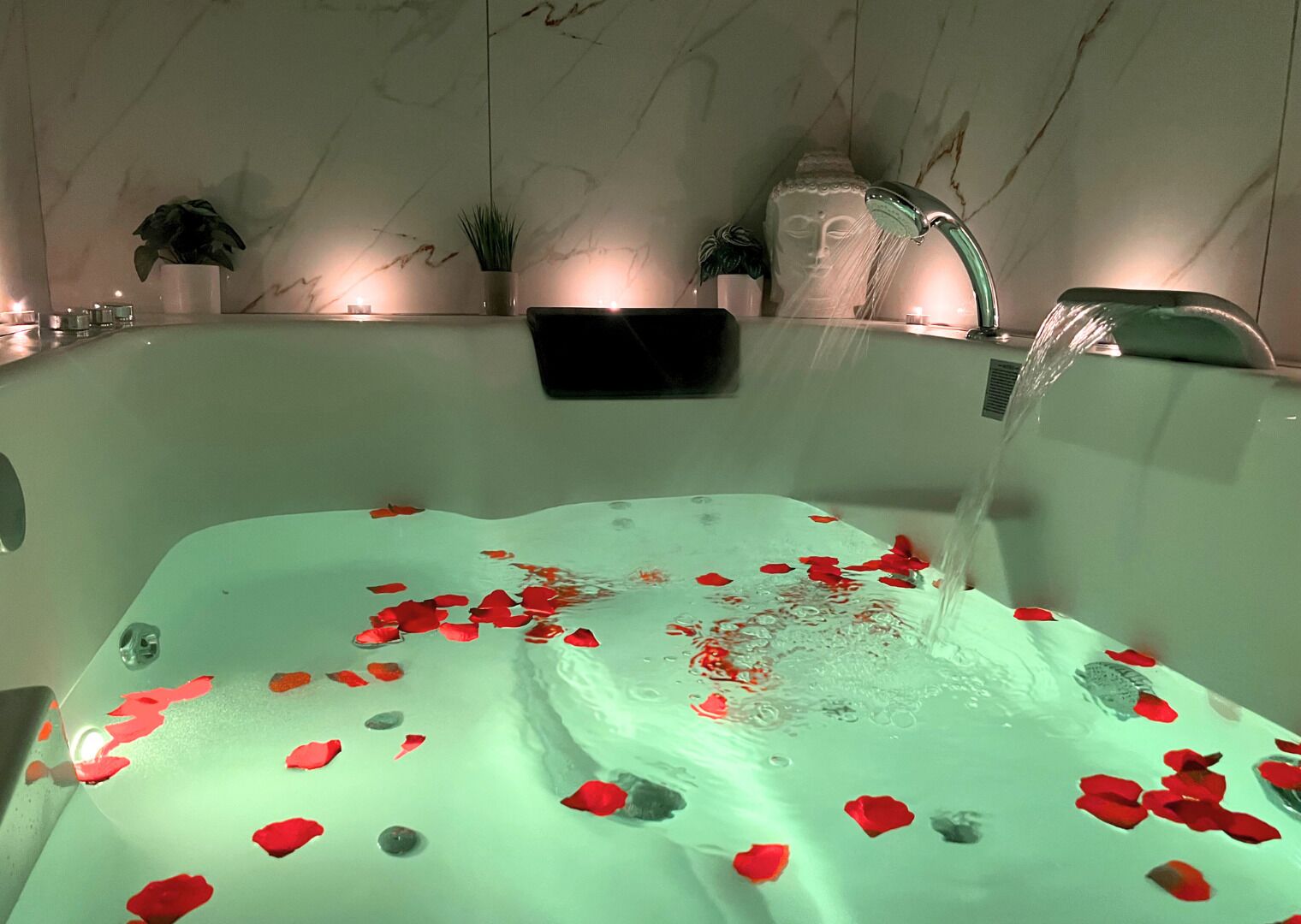 Eine mit Wasser und Rosenblättern gefüllte Badewanne, mit Marmorwand und einer Pflanze im Hintergrund.