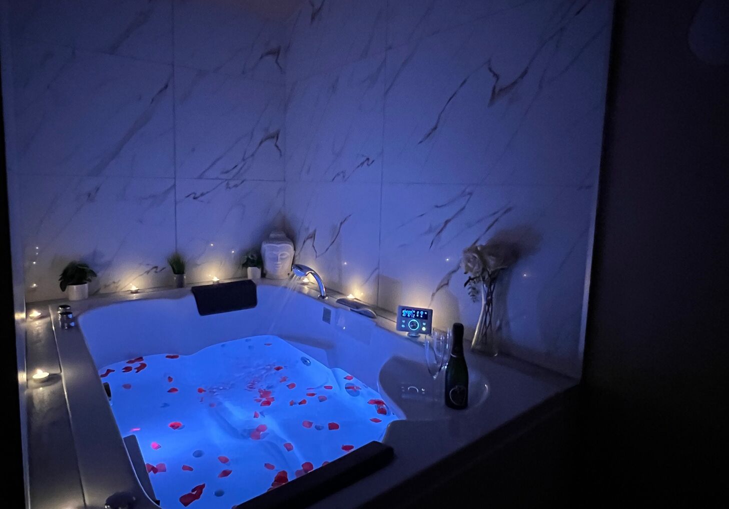 A modern bathroom with a marble wall, a hot tub with rose petals, a digital control panel, and a bottle of champagne.